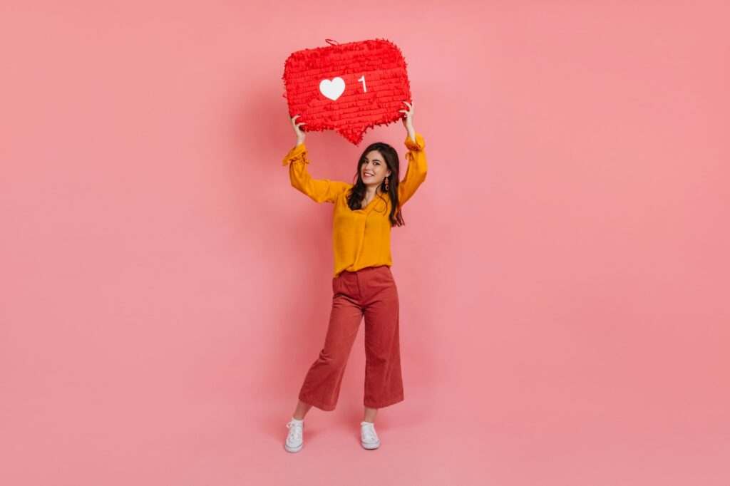 Full-length portrait of cheerful girl in culottes and yellow blouse holding “like” instagram si