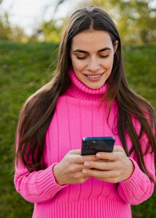 young smiling woman in pink sweater walking in green park using phone